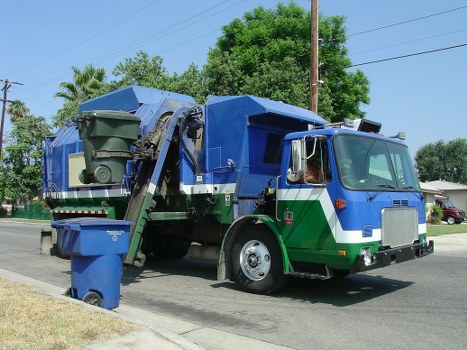 A large waste collection truck operated by Garden Clearance Muswell Hill parked on a residential street. The vehicle has a predominantly blue upper body with a green lower section, featuring a metallic finish and visible detailing such as the side mirrors, front grille, and headlights. The truck's rear arm is extended, lifting and tipping a green waste bin positioned at the back, indicating an active rubbish collection process. The bin is made of plastic with a textured surface and a hinged lid, currently open. The background shows a clear sky, utility poles with power lines, trees with lush green foliage, and parked cars along the curb. The scene reflects a typical rubbish removal service scene in a suburban area of Muswell Hill, with the environment well-maintained and the street quiet. This image illustrates the practical work of garden and rubbish clearance services, like those provided by Garden Clearance Muswell Hill, in a residential neighbourhood designed to manage and remove waste efficiently.