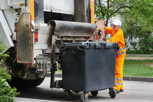 A waste collection operative dressed in high-visibility orange clothing and a white cap is standing beside a large black refuse bin with wheels, positioned on a paved street near a grassy area with trees. The operative is in the process of managing rubbish, possibly preparing for disposal or collection. The bin is made of plastic with a textured surface, and the wheels are visible at the base. The street environment includes a portion of a large waste collection vehicle with a metal compactor and hydraulic components on the left side of the image, indicating a professional rubbish removal service likely based in Muswell Hill. Surrounding greenery and a clear sky provide natural lighting, emphasizing the outdoor setting typical of local rubbish clearance operations by Garden Clearance Muswell Hill.