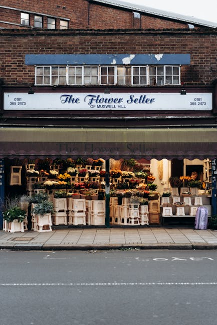 The image shows the storefront of a floral shop named 'The Flower Seller of Muswell Hill,' situated beneath a brick building. The shop features a large sign displaying its name and contact information